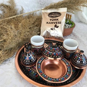 Puede incluir: Un juego de tres tazas de café turco tradicionales y platillos con diseños pintados a mano en tonos de azul, verde y rojo. Las tazas y los platillos están hechos de cobre y tienen una base negra. El juego se muestra en una bandeja de cobre con una base negra y un diseño similar pintado a mano. Una bolsa de café turco es visible en el fondo.
