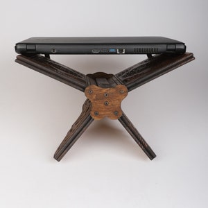 Op de afbeelding: Een donkerbruine houten laptopstandaard met ingewikkelde houtsnijwerk. De standaard is gevouwen in een driehoekige vorm, met een vlak oppervlak dat klaar is om een laptop te houden.