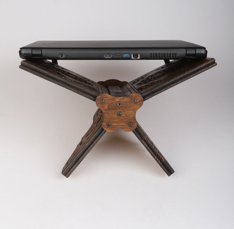 Op de afbeelding: Een donkerbruine houten laptopstandaard met ingewikkelde houtsnijwerk. De standaard is gevouwen in een driehoekige vorm, met een vlak oppervlak dat klaar is om een laptop te houden.