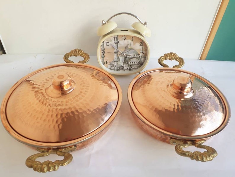 May include: Two copper serving dishes with a hammered finish and ornate gold handles. The dishes are round and have a domed lid.