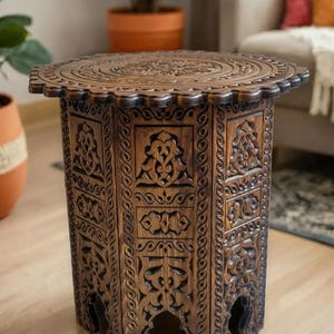 Può includere: Un tavolino da salotto in legno marrone scuro con intagli intricati. Il tavolo ha un piano rotondo e un bordo a festoni. Le gambe sono intagliate in un motivo decorativo.