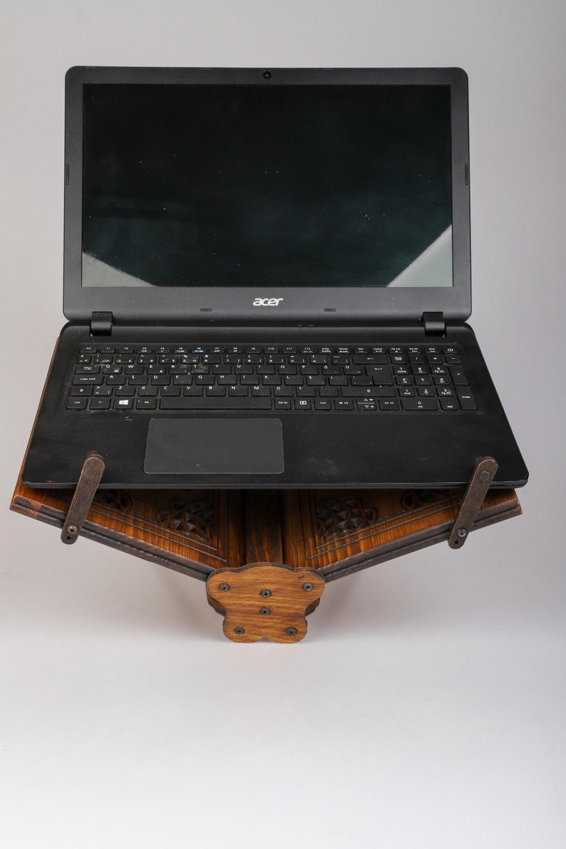 Op de afbeelding: Een zwarte laptop op een donkerhouten standaard. De standaard heeft ingewikkelde houtsnijwerk en is ontworpen om een laptop in een hoek te houden.