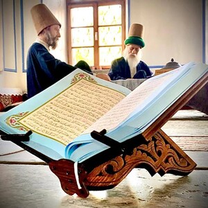 Op de afbeelding: Een houten boekenstandaard met ingewikkelde houtsnijwerk houdt een blauw boek met Arabische tekst vast. Twee mannen in traditionele hoeden zitten op de achtergrond.