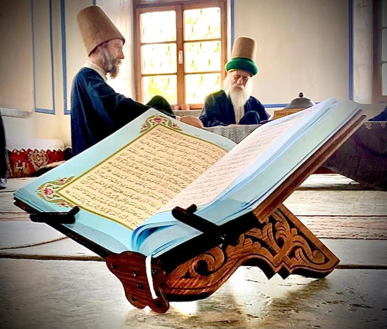 Op de afbeelding: Een houten boekenstandaard met ingewikkelde houtsnijwerk houdt een blauw boek met Arabische tekst vast. Twee mannen in traditionele hoeden zitten op de achtergrond.