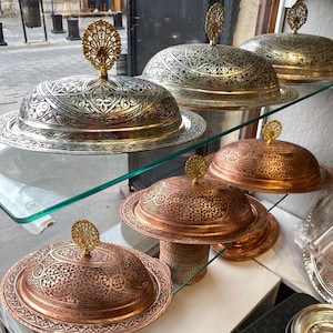 May include: A display of four ornate silver and copper serving dishes with intricate engraved designs. The dishes have domed lids with decorative finials.