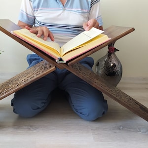 May include: A wooden prayer stand with intricate carvings, supporting an open book. The stand is in a light wood tone and has a cross-shaped design. The book is open to a page with yellowed paper and black text.