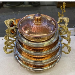 May include: A set of four copper and brass serving dishes with ornate handles. The dishes are stacked on top of each other, with the largest dish at the bottom and the smallest dish at the top. The dishes are all hammered and have a shiny finish.