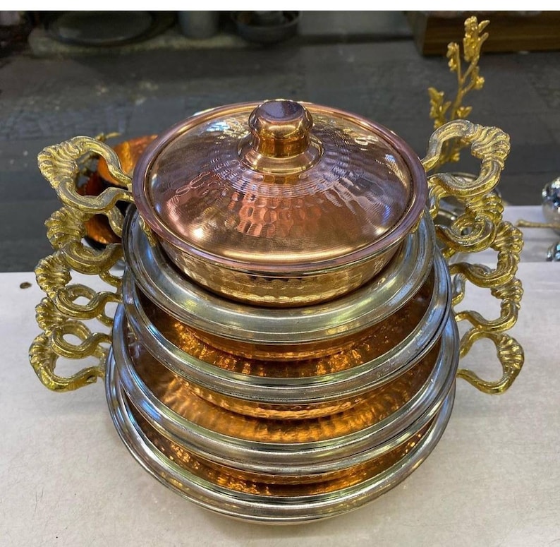 May include: A set of four copper and brass serving dishes with ornate handles. The dishes are stacked on top of each other, with the largest dish at the bottom and the smallest dish at the top. The dishes are all hammered and have a shiny finish.