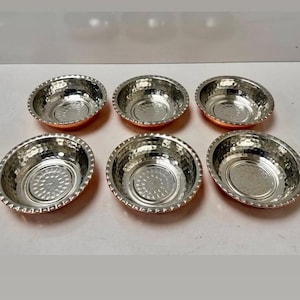 May include: Six small, hammered copper and silver-toned bowls. The bowls have a hammered texture on the exterior and a smooth, shiny interior. The bowls are arranged in a group on a white surface.