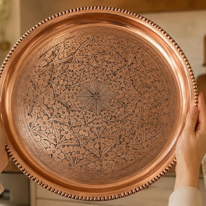 May include: A round copper tray featuring an intricate engraved floral pattern with a central star motif and a beaded rim. The metallic surface displays a warm, polished finish with dark etched details highlighting the botanical design elements.