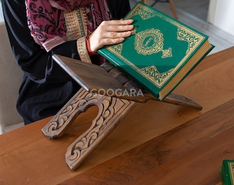 Op de afbeelding: Een houten Koranstandaard met sierlijke gravures ondersteunt een groen boek met gouden letters. Het boek is open en toont zijn pagina's. De standaard en het boek liggen op een houten oppervlak.