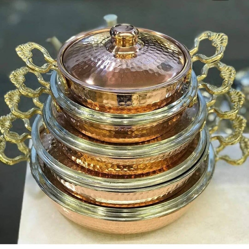 May include: A set of four copper pots with hammered finishes and gold-toned handles. The pots are stacked on top of each other, with the largest pot at the bottom and the smallest pot at the top. The pots have a shiny, metallic finish.