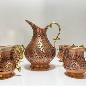 May include: A set of copper mugs and a pitcher with intricate floral designs. The mugs have gold handles and the pitcher has a gold spout. The set is perfect for serving beverages.