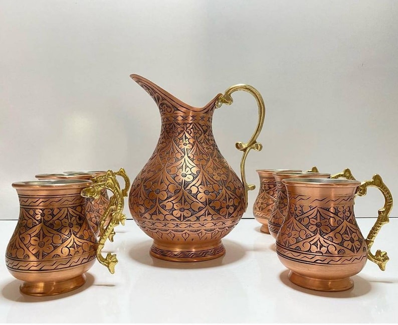 May include: A set of copper mugs and a pitcher with intricate floral designs. The mugs have gold handles and the pitcher has a gold spout. The set is perfect for serving beverages.