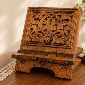Puede incluir: Un atril de madera marr&oacute;n con un intrincado patr&oacute;n tallado en el panel posterior y un borde con bisagras. La base incluye l&iacute;neas grabadas decorativas y peque&ntilde;os detalles de metal oscuro, proporcionando un soporte estable para los libros.