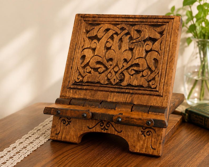 Puede incluir: Un atril de madera marr&oacute;n con un intrincado patr&oacute;n tallado en el panel posterior y un borde con bisagras. La base incluye l&iacute;neas grabadas decorativas y peque&ntilde;os detalles de metal oscuro, proporcionando un soporte estable para los libros.