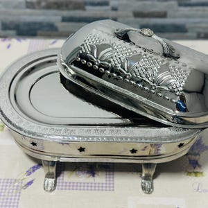 May include: A silver-colored, oval-shaped serving dish with a lid. The lid features a decorative floral design and a handle with a small gem. The dish sits on four ornate legs and has star-shaped cutouts.