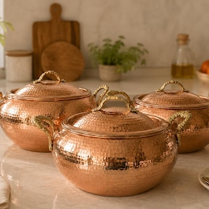 May include: A set of three round copper pots with hammered textures, each featuring a matching lid and two ornate, textured gold-toned metal handles on the sides. The pots have a polished metallic finish and are arranged in a cluster on a surface.