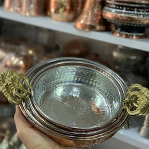 Op de afbeelding: Een set van drie koperen serveerschalen met gehammerde afwerkingen en goudkleurige handvatten. De schalen zijn in elkaar genest en hebben een glanzend, reflecterend oppervlak.