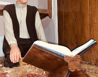 Op de afbeelding: Een man met een bruine hoed en een zwart vest zit op een tapijt met een houten boekenstandaard en houdt een open boek vast. De boekenstandaard heeft ingewikkelde houtsnijwerk en het tapijt heeft een rood, geel en blauw patroon.