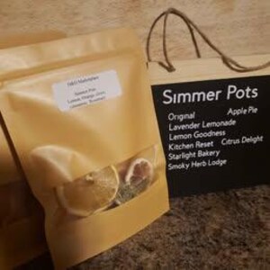 Peut inclure: Gros plan sur un sachet beige avec une fenêtre transparente, rempli de tranches d'agrumes séchées et d'herbes. Le sachet est à côté d'une enseigne en bois indiquant "Simmer Pots" et énumérant divers parfums.