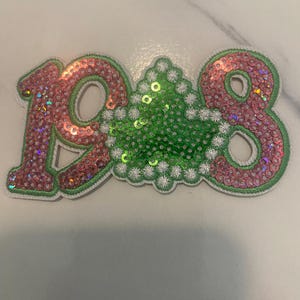 May include: A decorative patch featuring the numbers 1908 in pink sequins with a green sequined ivy leaf shape in the center. The numbers and leaf are outlined in white embroidery and green thread, creating a textured, layered appearance.