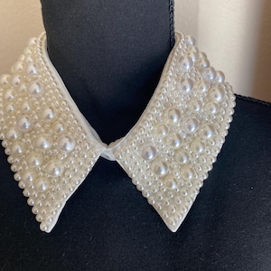 May include: A white, beaded collar with a classic design. The collar is adorned with numerous faux pearls of varying sizes, creating a textured, elegant appearance. The collar is displayed on a black mannequin.