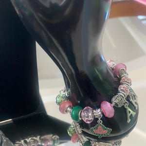 May include: A beaded bracelet with pink, green, and clear beads, and silver charms. The bracelet has a charm with a pink and green leaf design and the letter "A". The bracelet is displayed on a black surface.