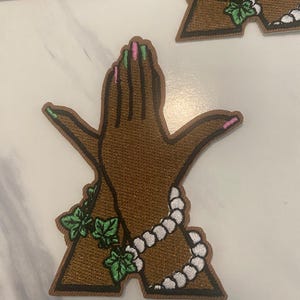 May include: An embroidered patch shaped like the letter A, featuring a brown hand with pink and green fingernails, wrapped in a white beaded strand and green ivy leaves. The design has a dark brown border and a textured, stitched surface.