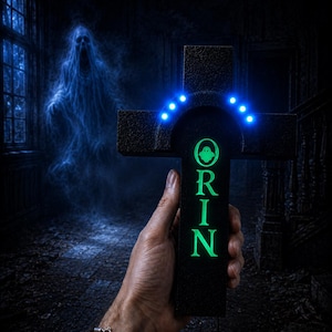 Op de afbeelding: Een zwart kruisvormig object met een getextureerd oppervlak, voorzien van een oplichtend groen verticaal opschrift dat ORIN leest met een klein spookicoon erboven, en een gebogen rij van zes helderblauwe LED-lampjes boven de tekst.