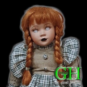 Op de afbeelding: Een vintage porseleinen pop met rood gevlochten haar, witte ogen en zwarte lippenstift. De pop draagt een geruite jurk en een bruin vest. De afbeelding bevat de tekst "devices for ghost hunting".