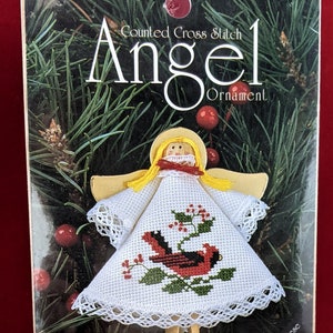 May include: A counted cross stitch angel ornament kit featuring a cardinal bird design. The kit includes a white fabric with a red cardinal bird, green holly leaves, and red berries. The angel is made of wood and has a yellow halo and blonde hair. The packaging is decorated with a green pine branch and red berries.