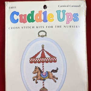 May include: A cross stitch kit for a nursery featuring a colorful carousel with a horse. The kit includes 18 count Aida fabric, presorted cotton floss, gold wrapped thread, a needle, instructions, and a frame. The kit is made in the USA.
