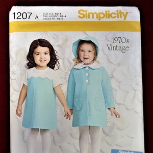 May include: Simplicity sewing pattern number 1207 for a vintage 1970s girls' dress and coat. The pattern is for sizes 1/2 to 4. The image shows two girls wearing the finished garments, one in a blue dress and the other in a blue coat with a white collar.