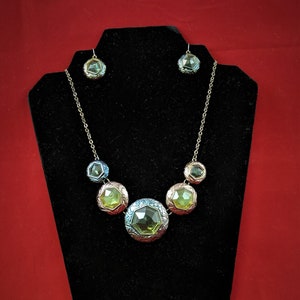 May include: A gold necklace with a graduated row of five faceted green and blue gemstones. The necklace is displayed on a black velvet stand. Matching earrings are also included.