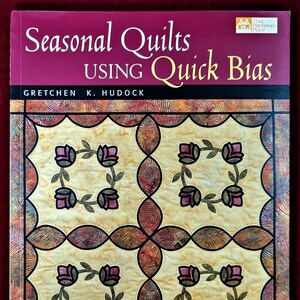 Könnte beinhalten: Buchcover von "Seasonal Quilts Using Quick Bias" von Gretchen K. Hudock. Es zeigt ein Quiltmuster mit vier floralen Quadraten in Rosa und Grün auf hellgelbem Hintergrund, umrahmt von einem dunkelroten Rand.