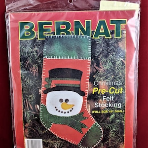 May include: A red and green felt stocking with a snowman face. The stocking is pre-cut and measures 18 inches in diameter. The package is labelled "Bernat Christmas Pre-Cut Felt Stocking (Full Size 18" Diag.) Stephanie's Collection."