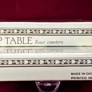 K&ouml;nnte beinhalten: Eine wei&szlig;e Schachtel mit goldenen Verzierungen und dem aufgedruckten Text "TOP TABLE Four coasters" auf der Oberseite. Die Unterseite der Schachtel tr&auml;gt den Text "MADE IN CHINA PRINTED IN HONG KONG".