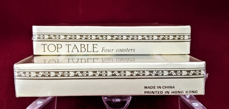 K&ouml;nnte beinhalten: Eine wei&szlig;e Schachtel mit goldenen Verzierungen und dem aufgedruckten Text "TOP TABLE Four coasters" auf der Oberseite. Die Unterseite der Schachtel tr&auml;gt den Text "MADE IN CHINA PRINTED IN HONG KONG".
