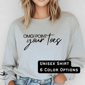 May include: A light gray unisex sweatshirt with the text "OMG! Point your toes" in black script. The sweatshirt has a crew neck and long sleeves. A black rectangle at the bottom says "UNISEX SHIRT" and "6 COLOR OPTIONS". A small logo is visible at the bottom left.