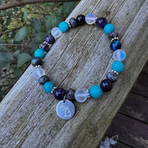 Peut inclure: Un bracelet de perles composé de perles rondes dans des tons turquoise, blanc translucide, violet foncé et gris moucheté, agrémenté de petites entretoises argentées et d'une breloque ronde argentée gravée du contour de l'État du Michigan.