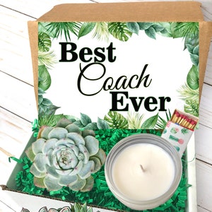 May include: A gift box with a succulent, candle, and matches. The box has the text "Best Coach Ever" and is decorated with tropical leaves. The succulent is a light green color, and the candle is in a clear glass jar.