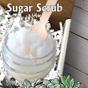 May include: A clear glass jar filled with white sugar scrub, with a wooden spoon resting inside. The jar sits in a cardboard box with white paper filler. The words "Sugar Scrub" are at the top. A banner with the text "Plants of Paradise" is at the bottom.
