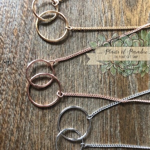 May include: Close-up of four necklaces with interlocking circle pendants. The necklaces are gold, rose gold, and silver, each with a delicate chain. The pendants are connected, creating a modern and minimalist design. The background is a wooden surface.