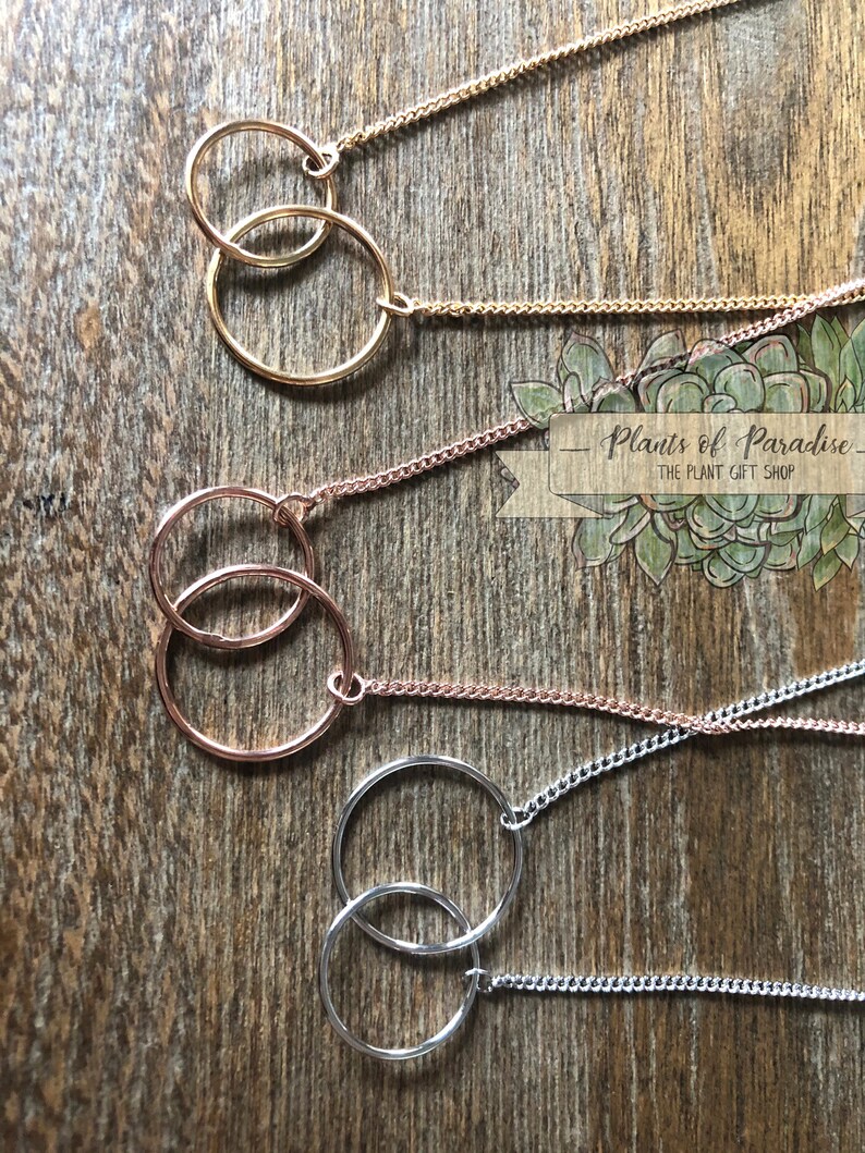 May include: Close-up of four necklaces with interlocking circle pendants. The necklaces are gold, rose gold, and silver, each with a delicate chain. The pendants are connected, creating a modern and minimalist design. The background is a wooden surface.