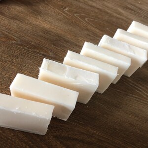 May include: Eight rectangular bars of white soap arranged diagonally on a wooden surface. The soap bars have a slightly uneven texture and are positioned to create a visual pattern. The wooden surface has a warm, brown tone.