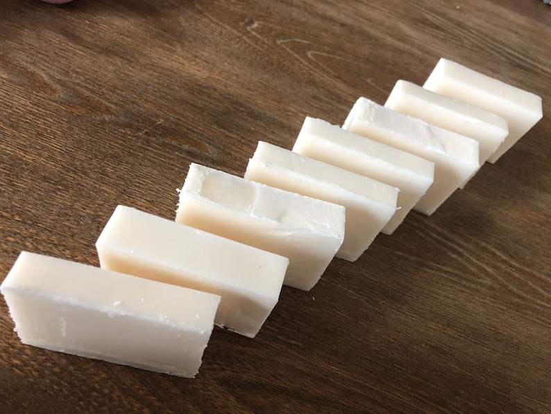 May include: Eight rectangular bars of white soap arranged diagonally on a wooden surface. The soap bars have a slightly uneven texture and are positioned to create a visual pattern. The wooden surface has a warm, brown tone.