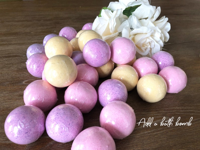 May include: A selection of bath bombs in pink, purple, and yellow, wrapped in clear cellophane. The bath bombs are arranged on a wooden surface, with white flowers in the background. Text reads "Add a bath bomb".