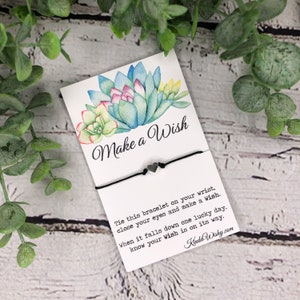 May include: A white card with a watercolor illustration of succulents and the text "Make a Wish". A black string bracelet with two silver heart charms is attached to the card. The card also includes instructions on how to use the bracelet to make a wish.