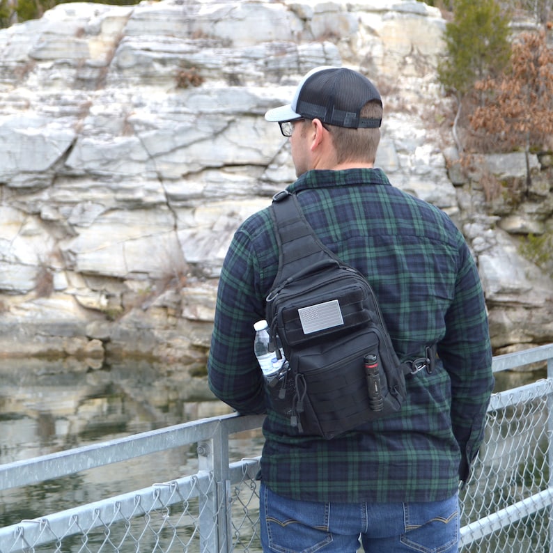 May include: A person wearing a black and green plaid shirt and a gray baseball cap is wearing a black tactical sling bag with a water bottle and a flashlight in the front pocket.